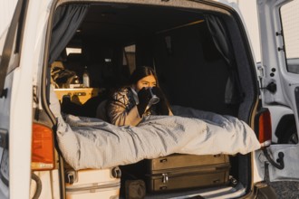 A Latina female relaxes with a coffee in a cozy van setup during a picturesque sunrise at Camp