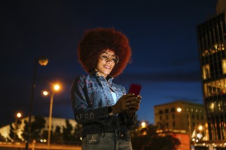 African descendent woman with afro hair, wearing glasses and a denim jacket, uses her smartphone on