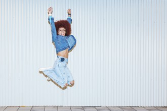 A joyful African descent woman with afro hair and fashionable denim outfit jumping energetically