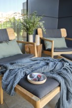 A stylish modern balcony featuring wooden chairs with cushions, a knitted throw, and a small table
