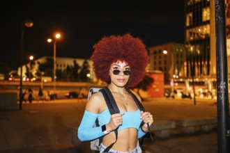 An African descendent woman with vibrant afro hair stands confidently on a city street at night.