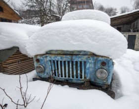 An old, rusty truck partially hidden beneath layers of thick snow in a rural winter setting in