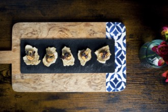 Crispy cracker canapes topped with vegetable satay stew and peanuts, arranged on a black slate over