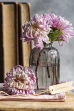 Soft pink chrysanthemums in a vintage glass vase beside rustic books and a tag reading Autumn