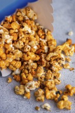 Caramel-coated popcorn spills from a blue container onto a textured gray surface, showcasing the