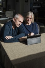 A senior couple sits at a table in a cozy living room, engaged with a digital tablet They appear to