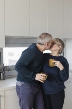 A senior couple embraces, sharing a tender kiss while holding coffee mugs in a bright, modern