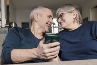 Smiling senior couple sitting on a couch, sharing a smartphone moment at home, demonstrating
