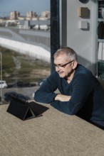 A senior man wearing glasses smiles as he engages with a tablet indoors Sunlight filters through