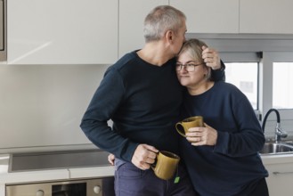 A senior couple shares a warm, affectionate moment in their kitchen, each holding a mug The scene