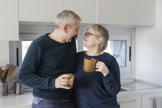 Senior couple shares a joyful moment in a modern kitchen, each holding a coffee cup Their warmth