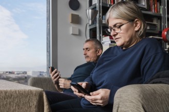 A senior couple sits in their cozy living room, each engaged with a smartphone The image captures