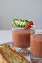 Clear glasses of gazpacho garnished with cucumber and tomato skewers A slice of crispy toast is