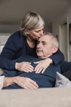 A senior couple shares a tender moment on the couch The woman lovingly kisses the man's head This