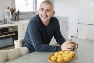 A cheerful senior man sits in a bright kitchen, holding a mug, with a basket of lemons nearby The