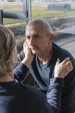 A senior couple shares a tender moment near a sunlit window, with one gently holding the other's
