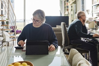 A senior couple sits in a bright home office, one using a tablet, the other focusing on a computer