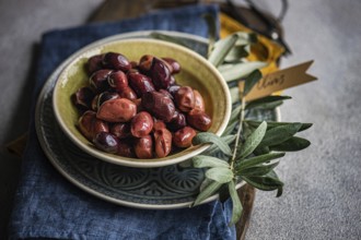 Bowl of vibrant Greek olives, accompanied by a sprig of fresh olive leaves, in a blue napkin. with