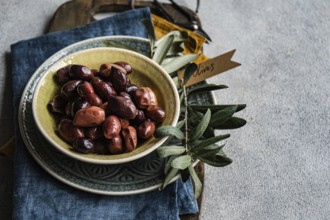 Ceramic bowl filled with Greek BBQ olives, accompanied by a strand of fresh olive leaves and a
