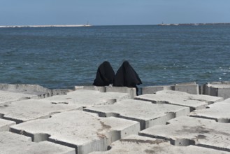Two Muslim women wearing black abaya or chador sit on the concrete breakwater on the Mediterranean