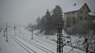 Snowy landscape with railway line, overhead lines and house, surrounded by trees under grey skies,