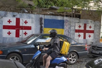 Tbilisi, Georgia. August 26th 2025. A motorcycle delivery driver rides past the flags of Georgia