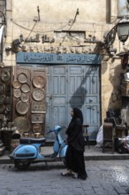 Cairo, Egypt. April 15th 2025. A Muslim woman walks past a blue motor scooter and antique shop in