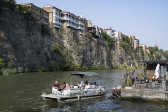 Tbilisi, Georgia. July 29th 2025. A tourist river cruise passes under building sitting precariously