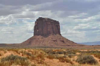 Monumental rock rises from sandy desert under cloudy sky, Monument Valley, Navajo Tribal Park,