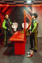 Two young men in green and black jumpsuits chat in a red neon locker room, gearing up and bonding