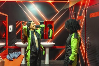 Two young women in neon lit locker room laughing and suiting up in racing jumpsuits, bonding before