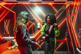 Two cheerful women are preparing for a gokarting race in a neon lit locker room, with one friend