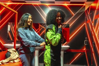 Two diverse friends laughing and helping each other put on racing suits in a changing room with