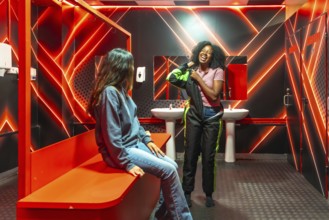Two diverse young women laughing and dressing in race suits inside a modern changing room with red