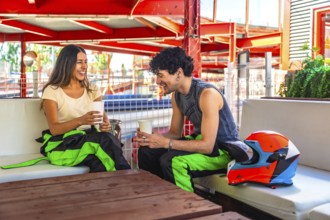 Young couple in racing suits sharing coffee and laughter at a trackside table after karting,