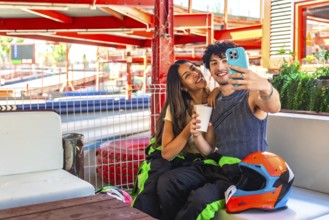 Young couple wearing racing suits sitting on a bench at a go kart circuit, smiling and taking a