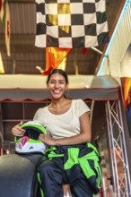 Young woman in racing suit sits in a go kart holding her helmet beneath a checkered flag, smiling