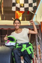 Young woman smiling and raising a fist in triumph, holding a karting helmet, wearing a racing suit,