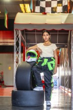 Woman wearing a racing suit and helmet standing confidently by tires and checkered flags at an