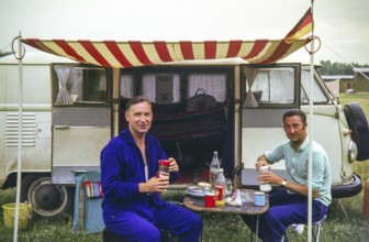 Two men having breakfast at campsite sitting outside a VW Volkswagen camper van vehicle, 1971
