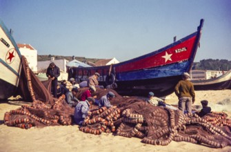 Fishermen repairing nets on beach by traditional fishing boats, Nazaré, Portugal, Europe 1968