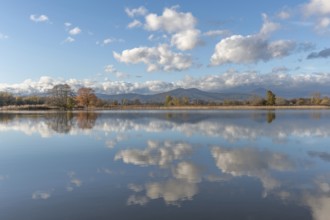 The river flows flat and reflects clouds and mountains under a clear sky. Autumn trees draw the