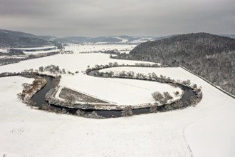 River flows in a loop through snow-covered fields, framed by hills and trees under cloudy skies,