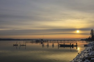 A jetty stretches into a calm lake at sunrise, boats rest on the shore, the sky is yellow and gray,