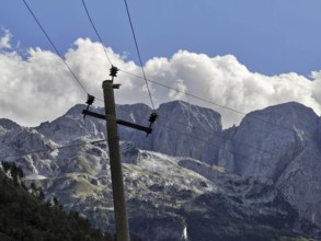 Leaning wooden power poles against rocky mountains and imposing clouds in a bright blue sky, Peaks