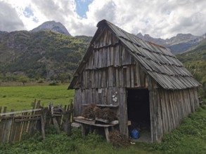 Rustic wooden cabin in a green meadow against an alpine mountain backdrop with cloudy sky, Peaks of