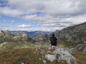 Person with backpack sitting on a rock and looking at a mountain panorama under cloudy sky, Peaks