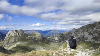 Hiker with backpack enjoying the view of a wide mountain valley under a partly cloudy sky, looking