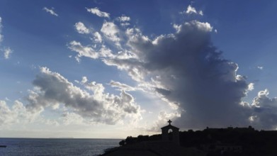 Dramatic sky with clouds and sunlight over sea with church silhouette in foreground, Himare,