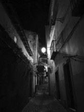 A narrow alley at night with illuminated alleyway lamp between tall, dark buildings, monochrome,